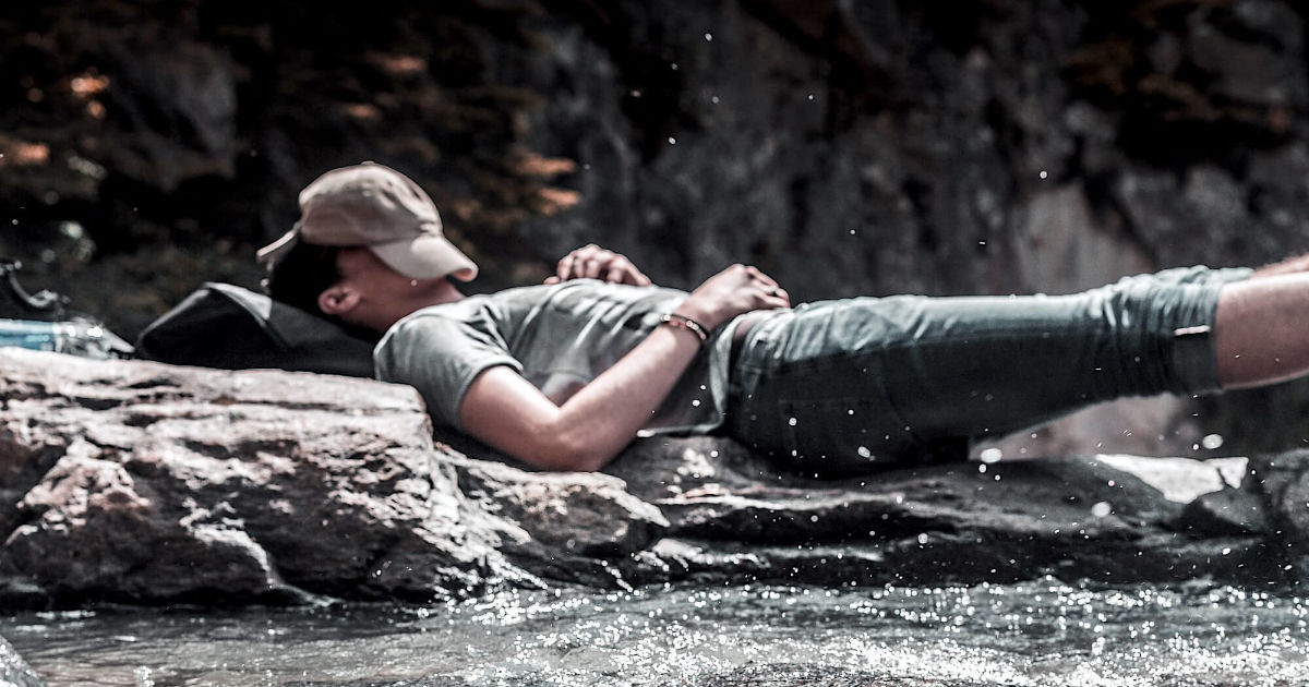 Мужчина спит на берегу горной реки, укрыв лицо кепкой
