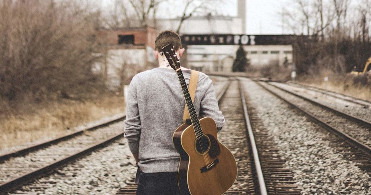 Парень с гитарой за спиной стоит на заброшенных железнодорожных путях