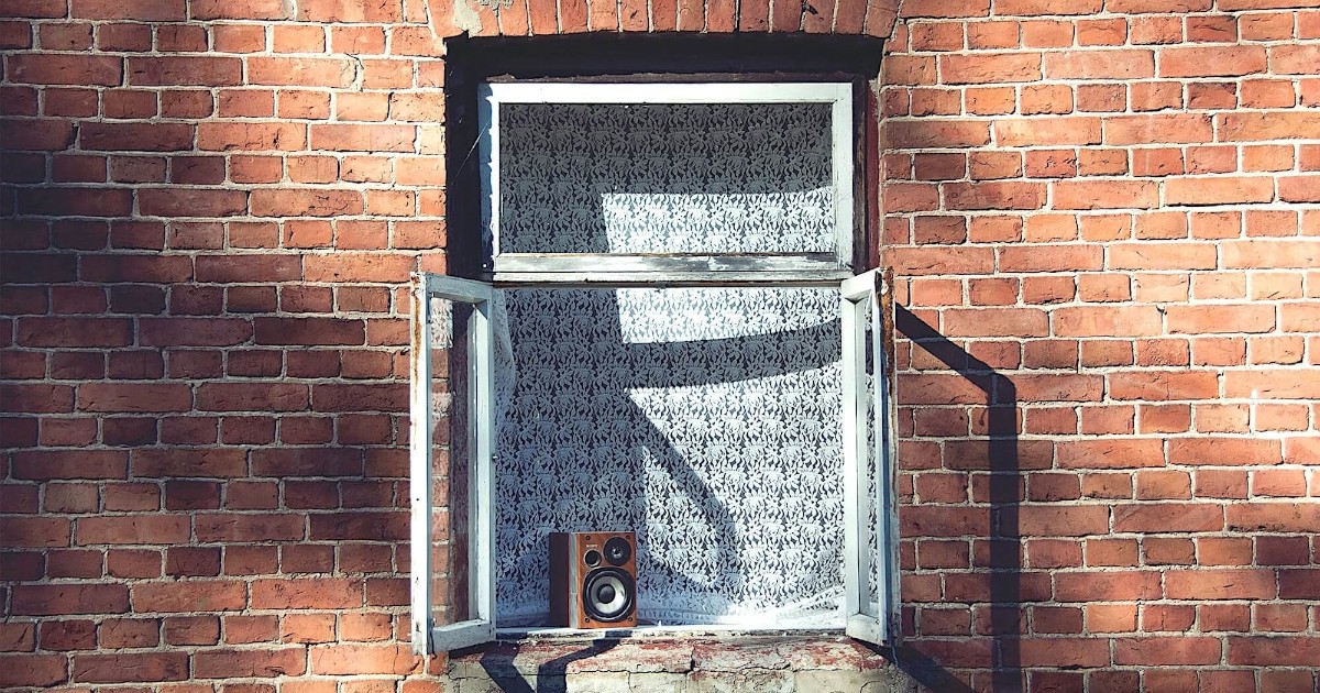 Полочная акустика в виде одной колонки, стоящей на подоконнике раскрытого окна дома из красного кирпича