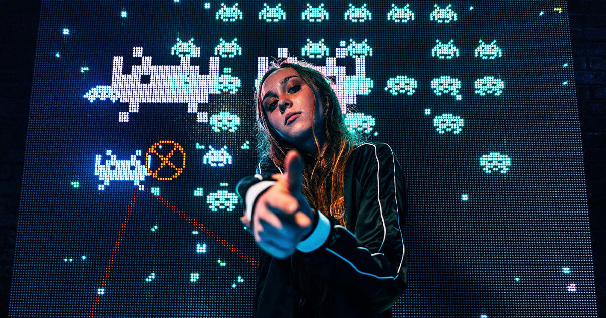 Девушка-геймер направляет вперёд руку со стреляющим жестом, на фоне экрана с восьмибитной игрой