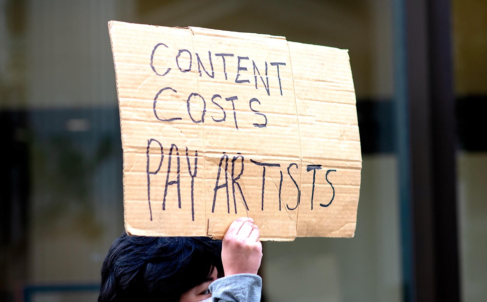 Азиатская женщина держит над собой картонный плакат с надписью на английском «Контент стоит денег – платите деятелям искусства»