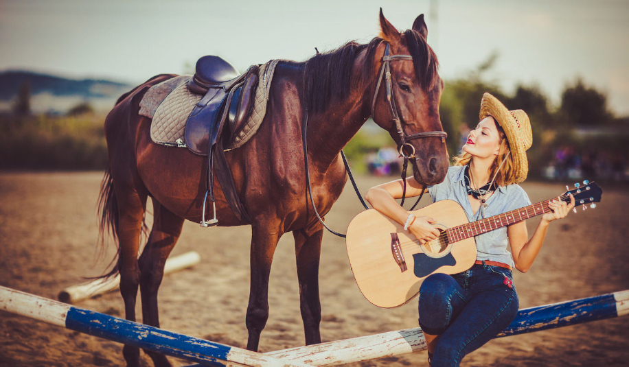 Женщина в ковбойской шляпе играет на акустической гитаре рядом с лошадью, сидя на манеже для конкура